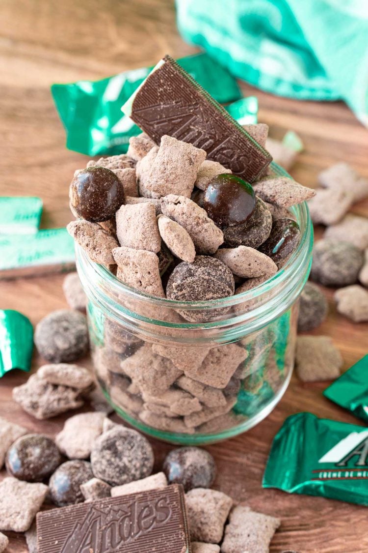 Mint chocolate muddy buddies in a jar on a wooden table surrounded my muddy buddies and mint candies.