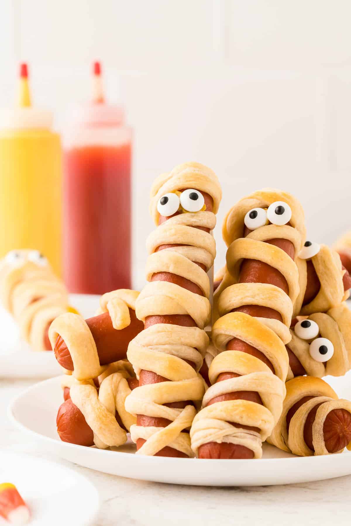 Mummy hot dogs on a white plate with mustard and ketchup in the background.