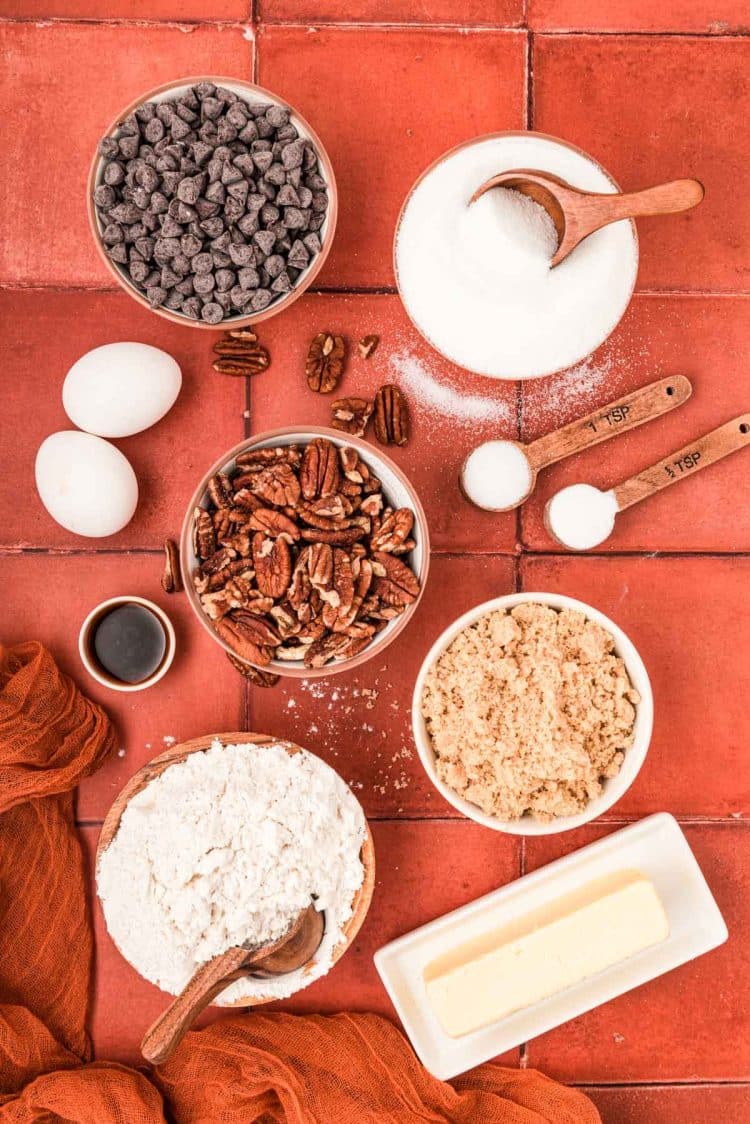 Ingredients to make pecan chocolate chip cookies on a rust colored tile surface.