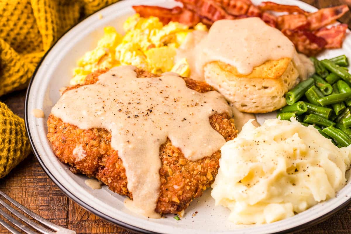 Country friend steak on a white plate with mashed potatoes, eggs, bacon, and a biscuit.