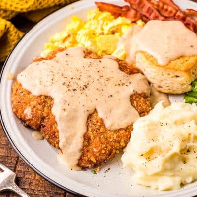 Country friend steak on a white plate with mashed potatoes, eggs, bacon, and a biscuit.