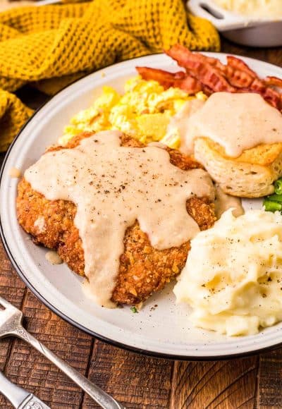 Country friend steak on a white plate with mashed potatoes, eggs, bacon, and a biscuit.