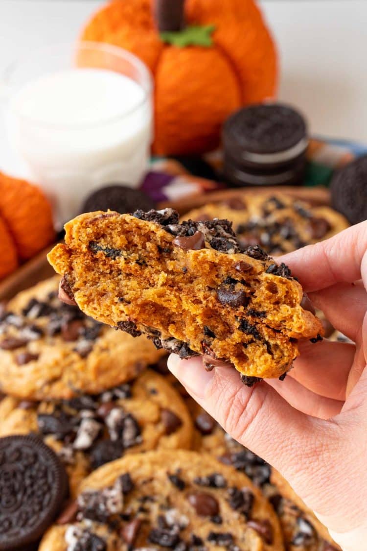 A woman's hand holding a pumpkin chocolate chip cookie with oreos in it broken in half so you can see the chewy center.