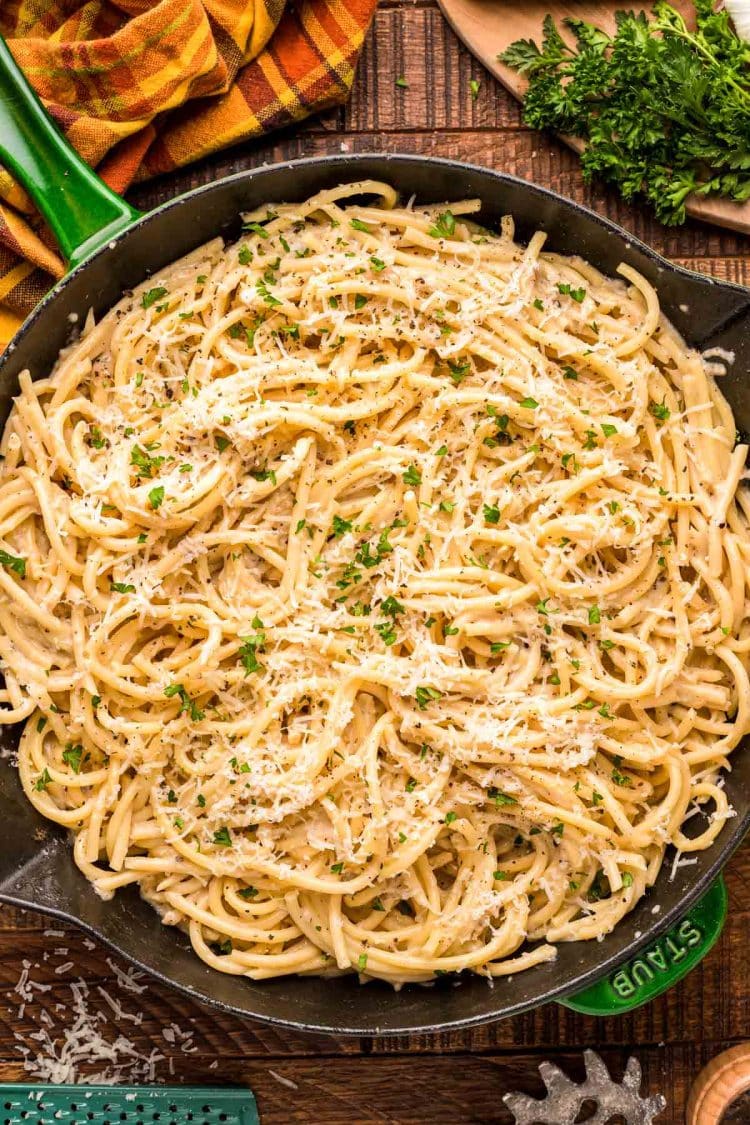 Overhead photo of bucatini cacio e pepe in a green and black skillet on a wooden table.