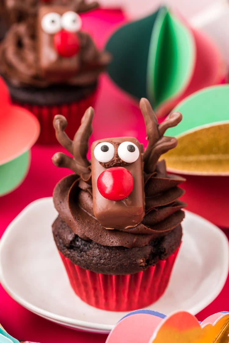 Reindeer decorated chocolate cupcake on a white plate with holiday decorations in the background.