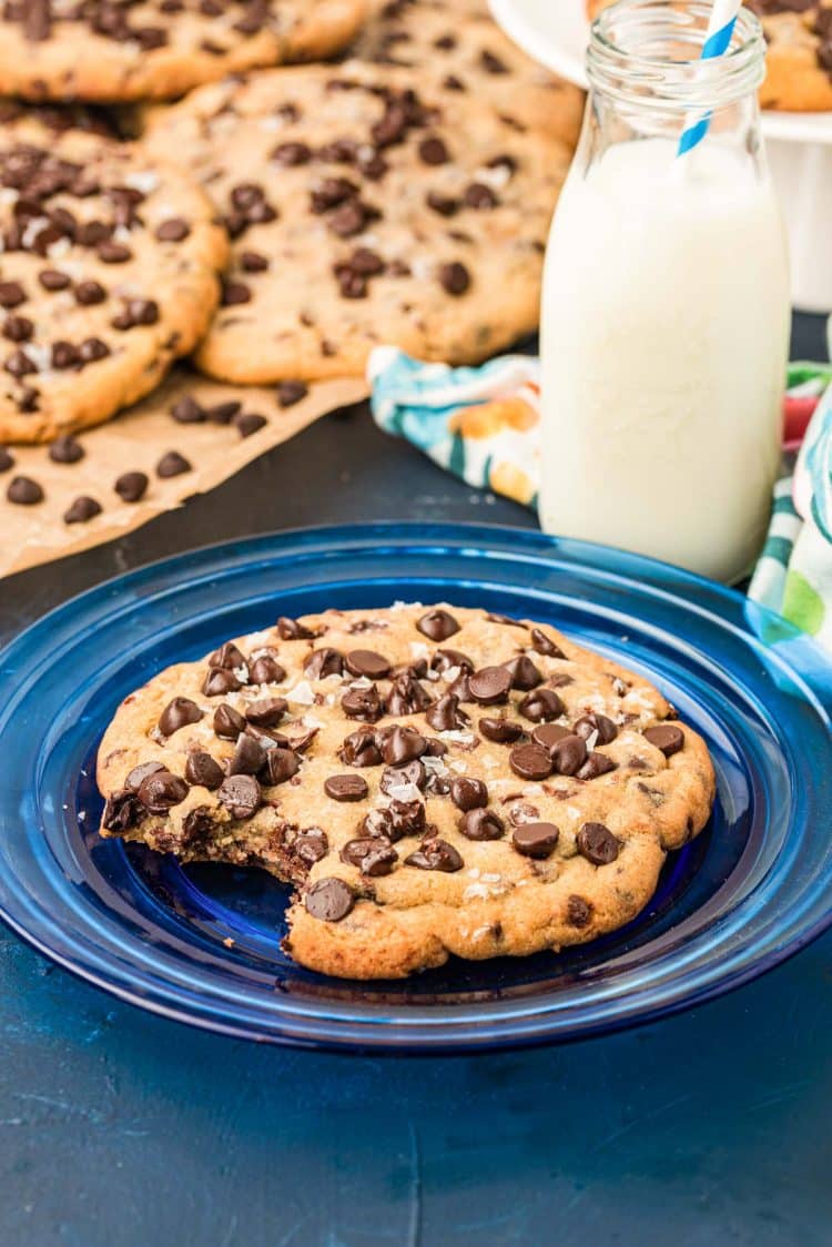 A giant chocolate chip cookie with a bite taken out of it on a blue plate with a bottle of milk next to it and more cookies in the background.