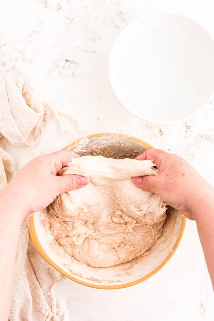 A woman's hand reaching under a ball of sourdough dough in a bowl.