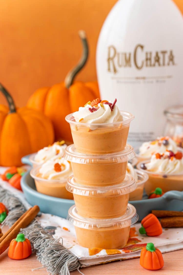 A stack of four pumpkin pie pudding shots with a bottle of rumchata and pumpkins in the background.
