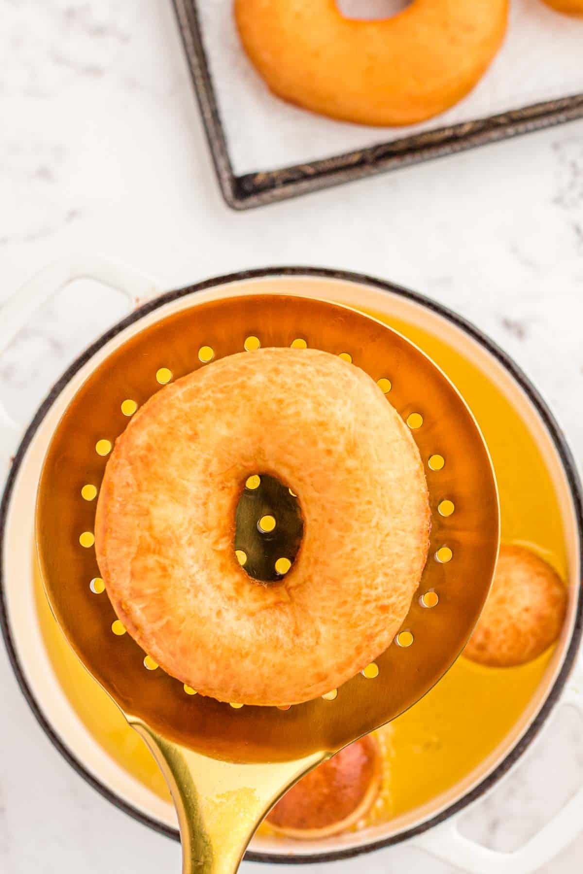 A slotted spoon lifting a fried donut out of oil pan.