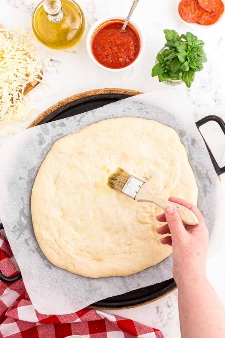 Sourdough pizza crust being shaped and brushed with olive oil on a parchment lined-pizza stone.