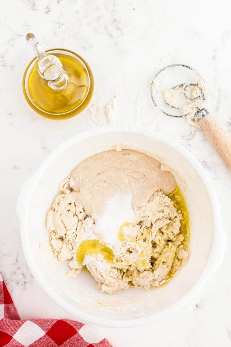 Oil, salt, and sourdough starter being added to a bowl with dough to make pizza crust.