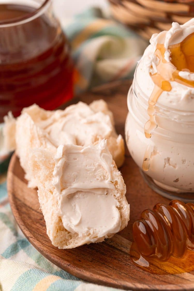 Dinner rolls halves slathered with whipped honey butter next to a jar of it on a wooden plate.