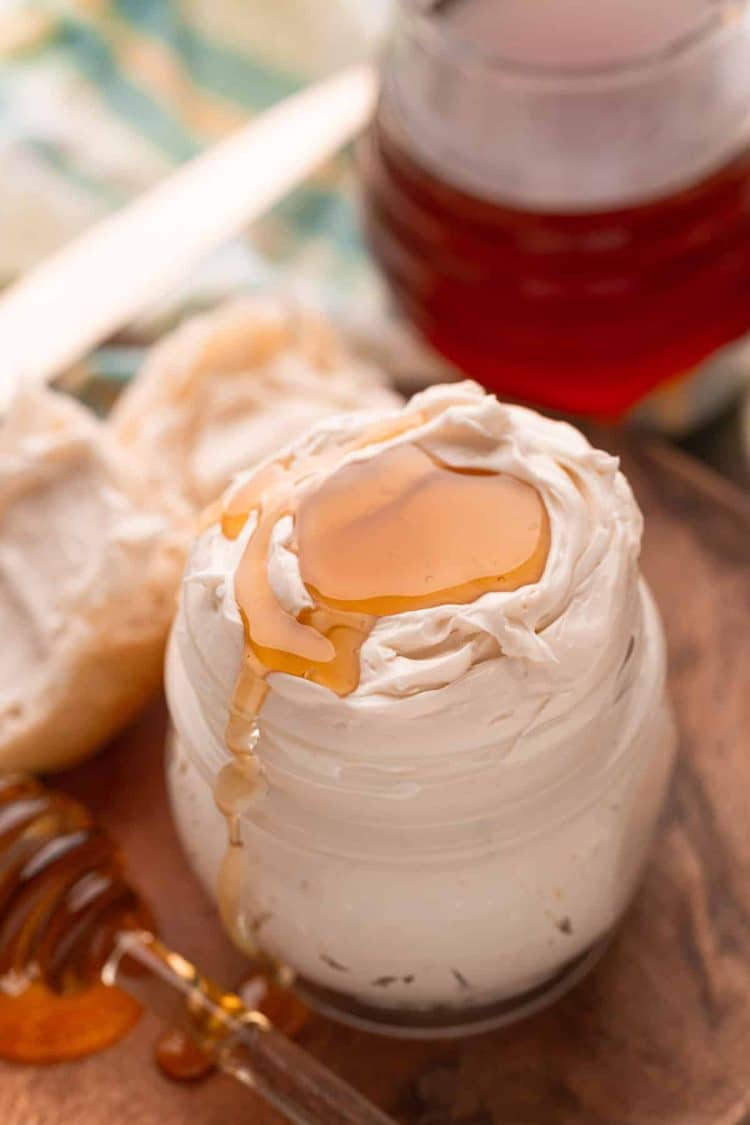 Honey butter in a glass jar on a wooden plate with a roll next to it.