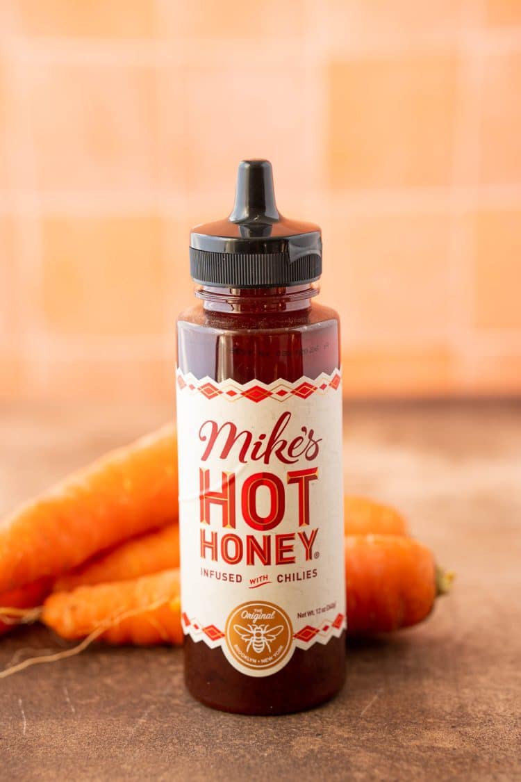 A bottle of hot honey on a table with carrots behind it.