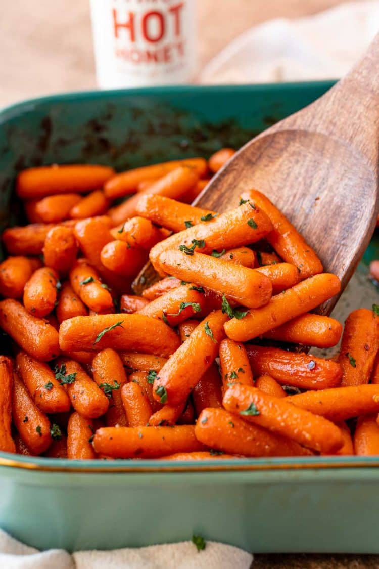 A serving spoon scooping hot honey glazed carrots out of a baking dish.