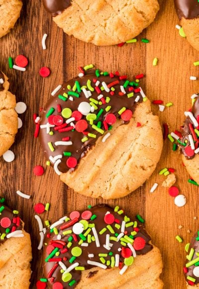 Peanut butter cookies dipped halfway in chocolate and coated with holiday sprinkles on a wooden board.