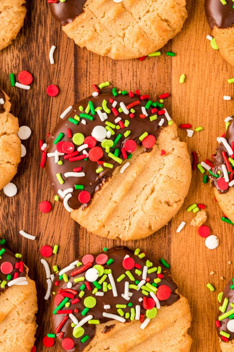 Peanut butter cookies dipped halfway in chocolate and coated with holiday sprinkles on a wooden board.