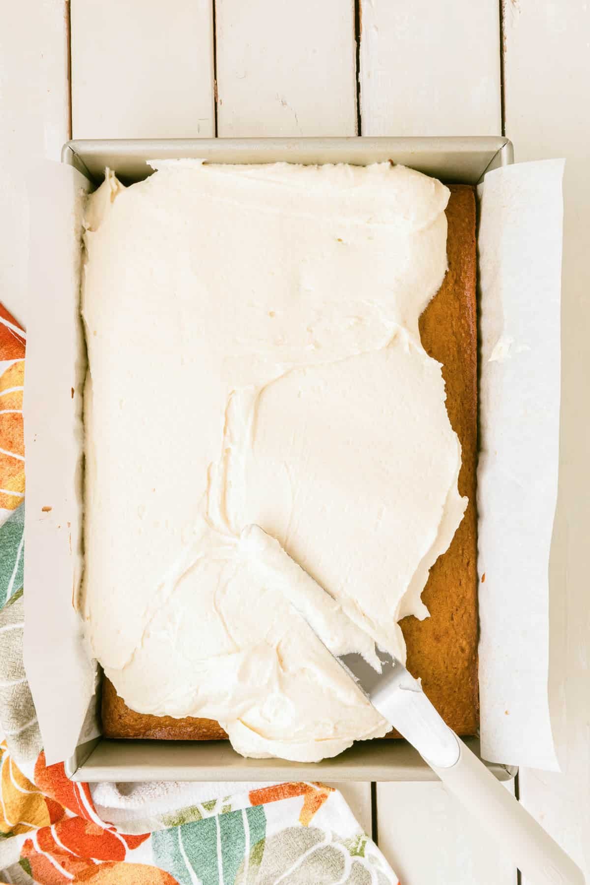Cream cheese frosting being spread over a pumpkin cake in a pan.
