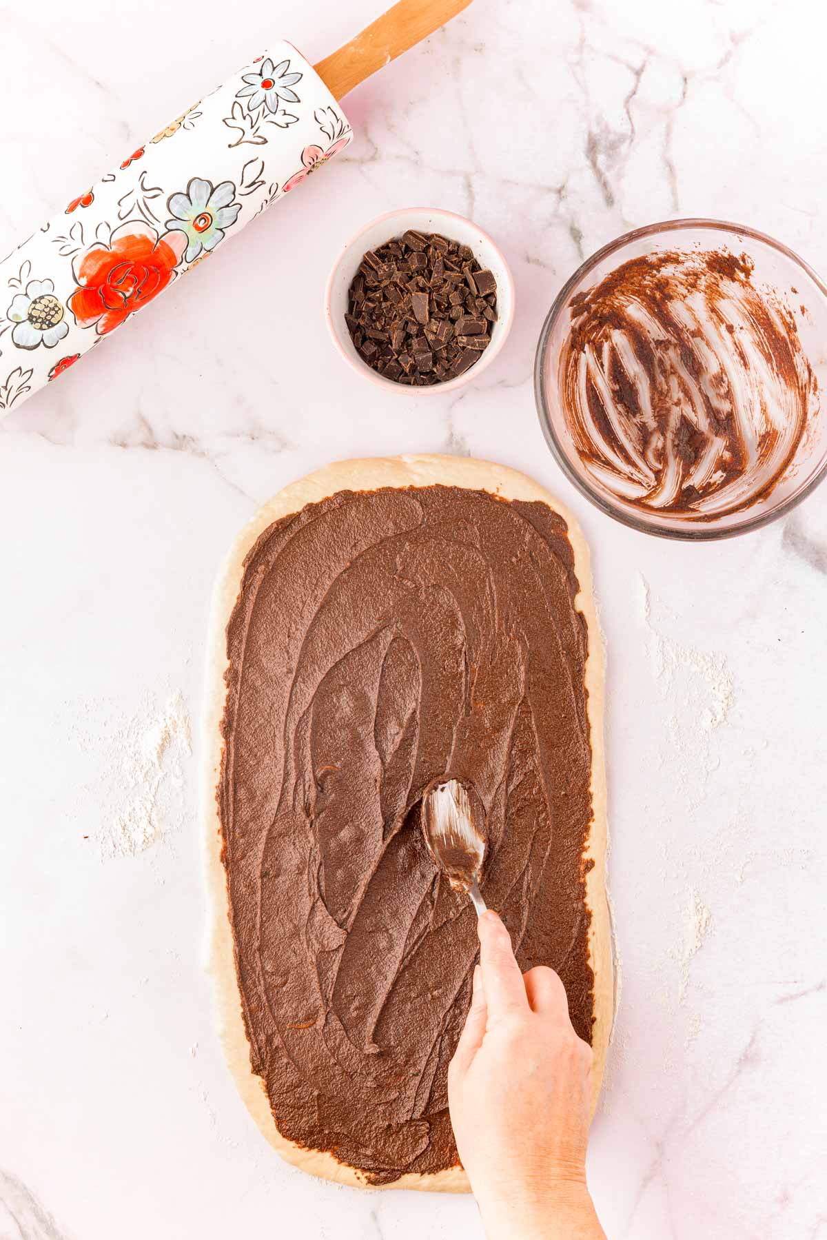 A woman using a spoon to spread chocolate cinnamon roll filling on a dough to make cinnamon rolls.