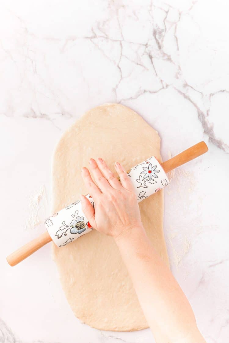 A woman's hand using a rolling pin to shape dough for cinnamon rolls.