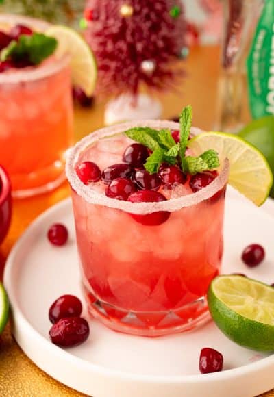 Close up of a Cranberry Margarita on a white tray on a gold surface with limes and cranberries around it.