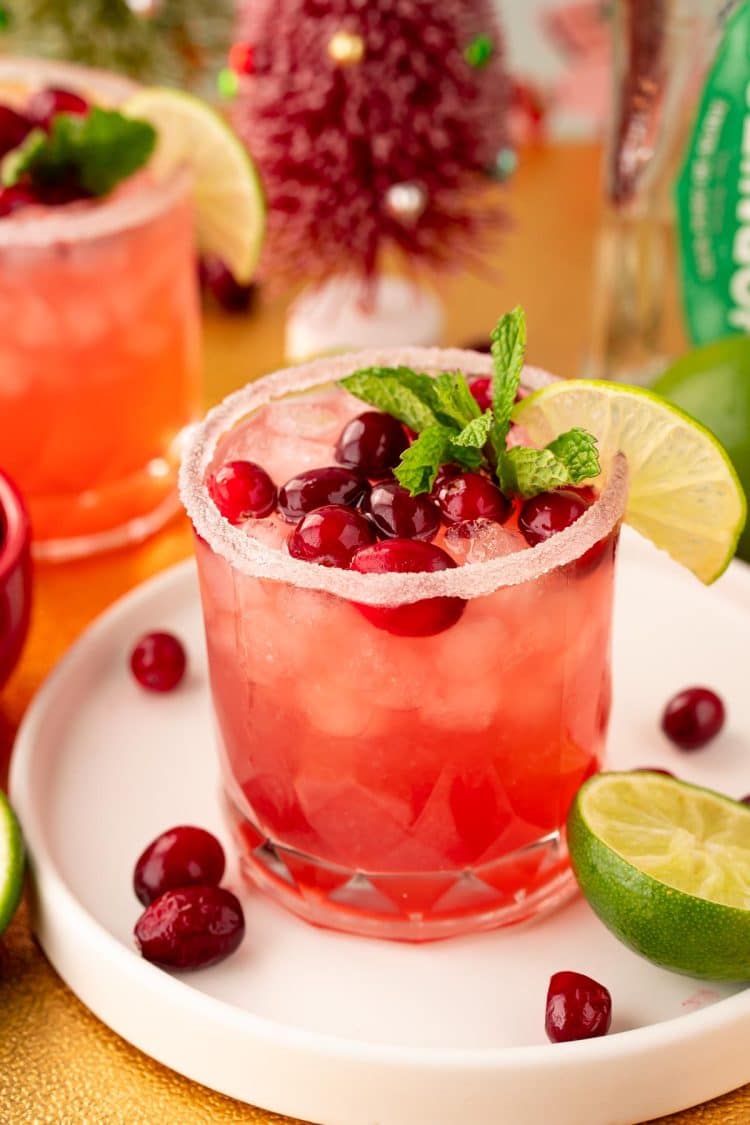 Close up of a Cranberry Margarita on a white tray on a gold surface with limes and cranberries around it.