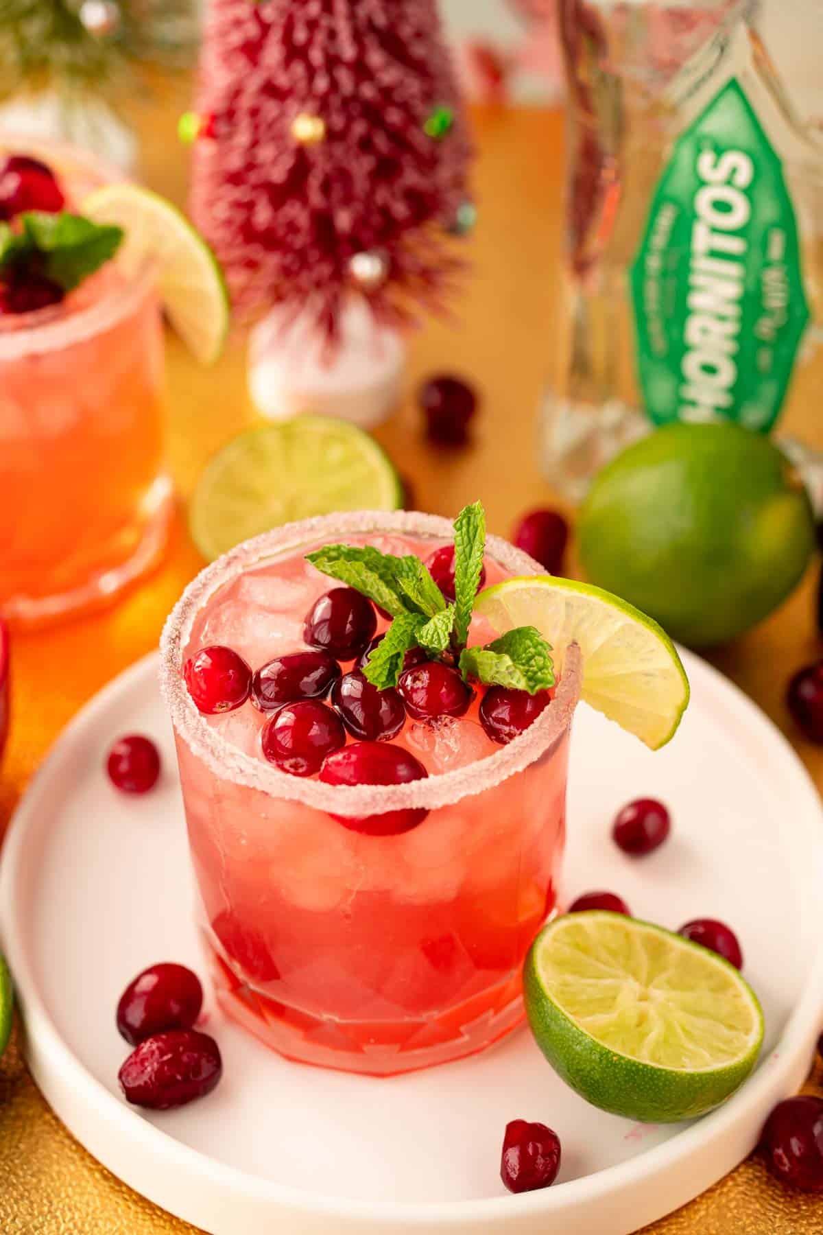 A Chritstmas Cranberry margarita on a white serving tray surrounded by limes and cranberries.
