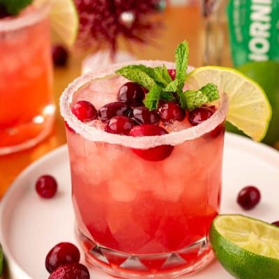Close up of a Cranberry Margarita on a white tray on a gold surface with limes and cranberries around it.