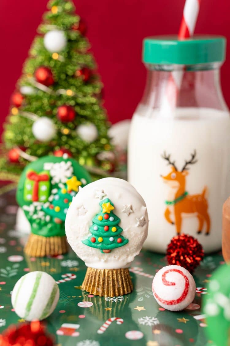An Oreo Snow Globe Cookie with a Christmas tree on it with more decorations and cookies and milk in the background.