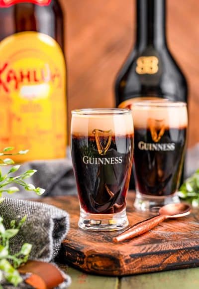 Baby Guinness shots in little Guinness shot glasses on a wooden serving board with bottles or Bailey's and Kahlua in the background.