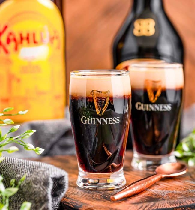 Baby Guinness shots in little Guinness shot glasses on a wooden serving board with bottles or Bailey's and Kahlua in the background.