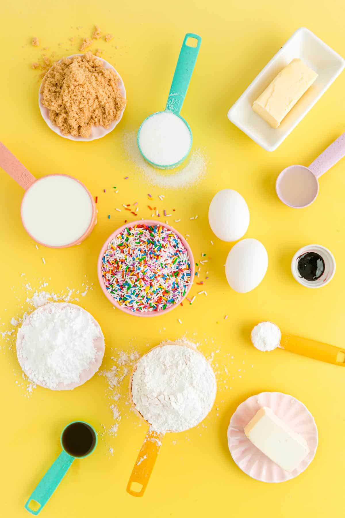 Ingredients to make birthday cake donuts on a yellow surface.
