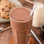A tall glass of chocolate milk on a leather coaster on a brown counter with milk and cookies in the background.
