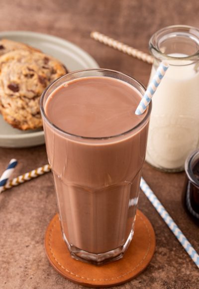 A tall glass of chocolate milk on a leather coaster on a brown counter with milk and cookies in the background.