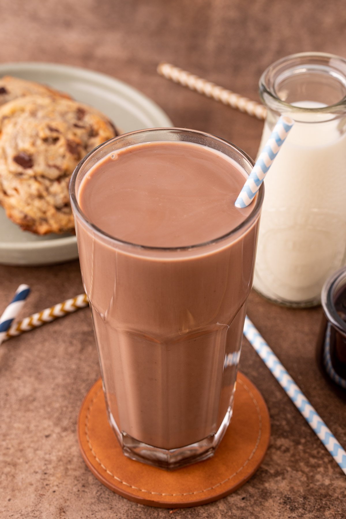 A tall glass of chocolate milk on a leather coaster on a brown counter with milk and cookies in the background.