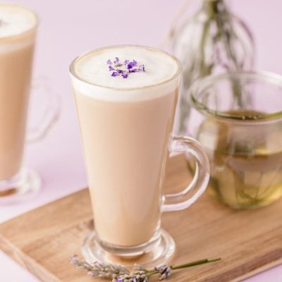 Two mugs of lavender lattes on a wooden serving board on a purple surface.