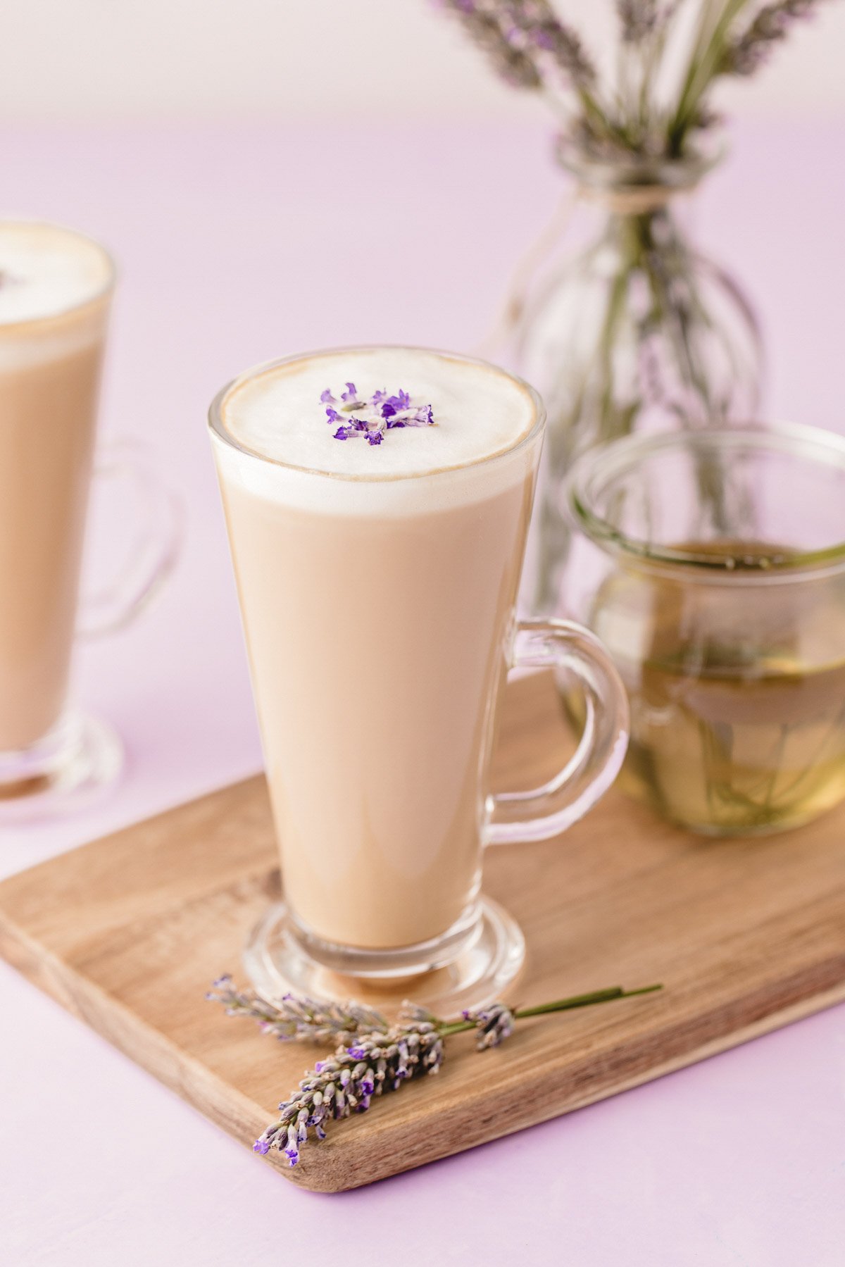 Two mugs of lavender lattes on a wooden serving board on a purple surface.