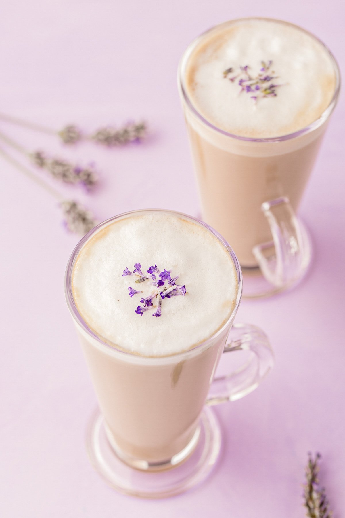 Two lavender lattes in glass mugs on a purple surface.