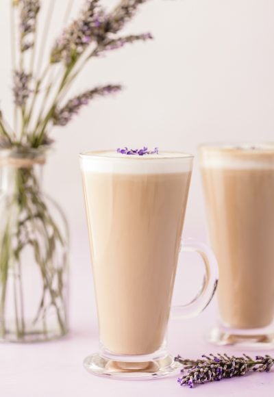 A tapered glass mug with a lavender latte in it, another in the background.