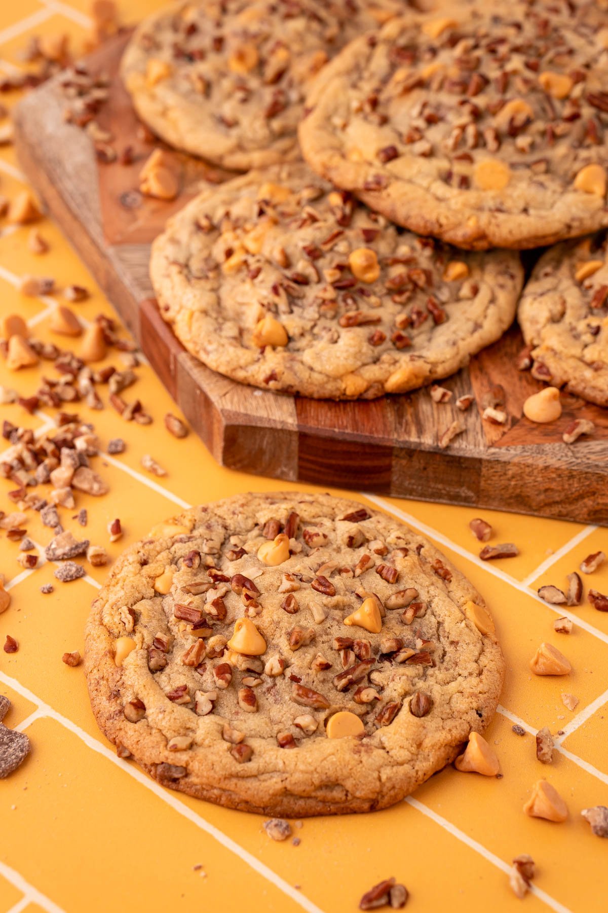 Butter pecan cookies on a yellow table, some on a serving board with pecans and butterscotch chips scattered around it.