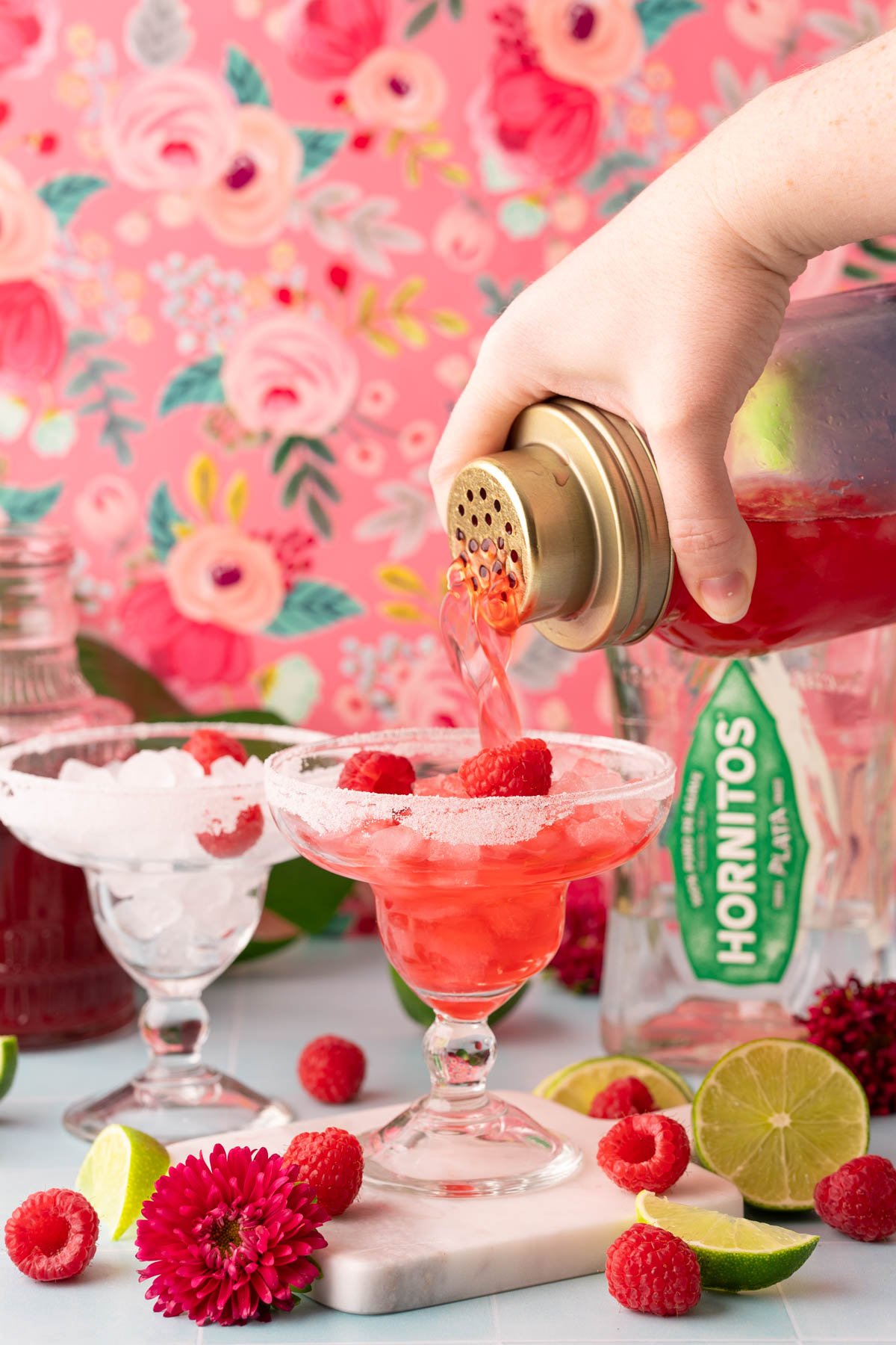 A cocktail shaker straining a raspberry margarita into margarita glasses.