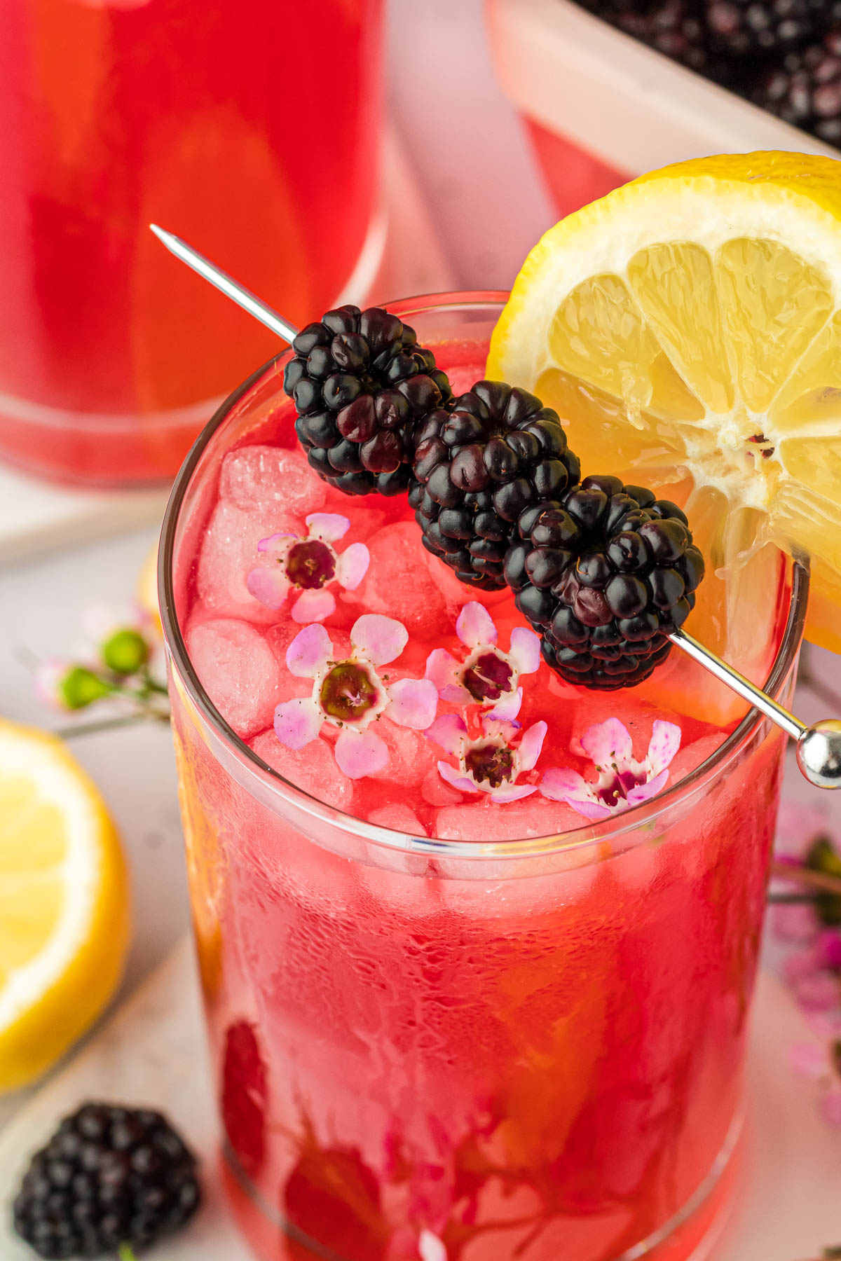 Fresh blackberry lemonade in a glass garnished with lemon, blackberries, and flowers.