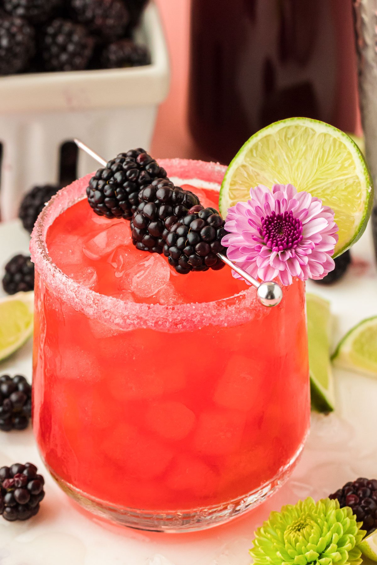 Close up photo of a blackberry margarita on a table garnished with blackberries, lime, and a flower.