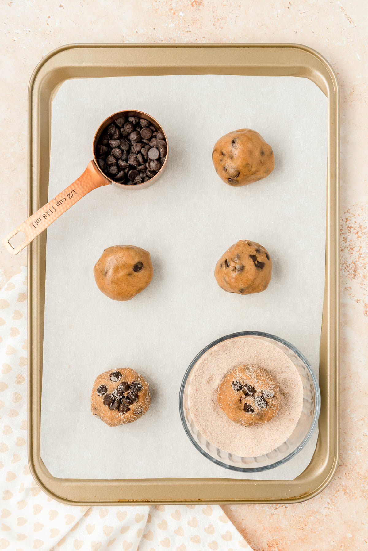 Balls of cinnamon chocolate chip cookie dough being rolled in cinnamon sugar on a baking pan.