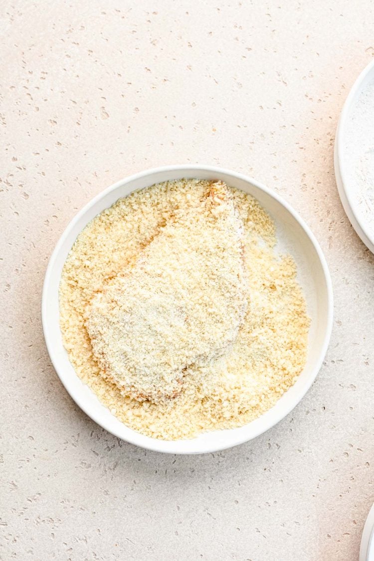 Chicken being coated in panko bread crumbs in a white bowl.