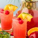 A glass of fruit punch on a wooden coaster with another glass, pitcher, and fruit in the background.