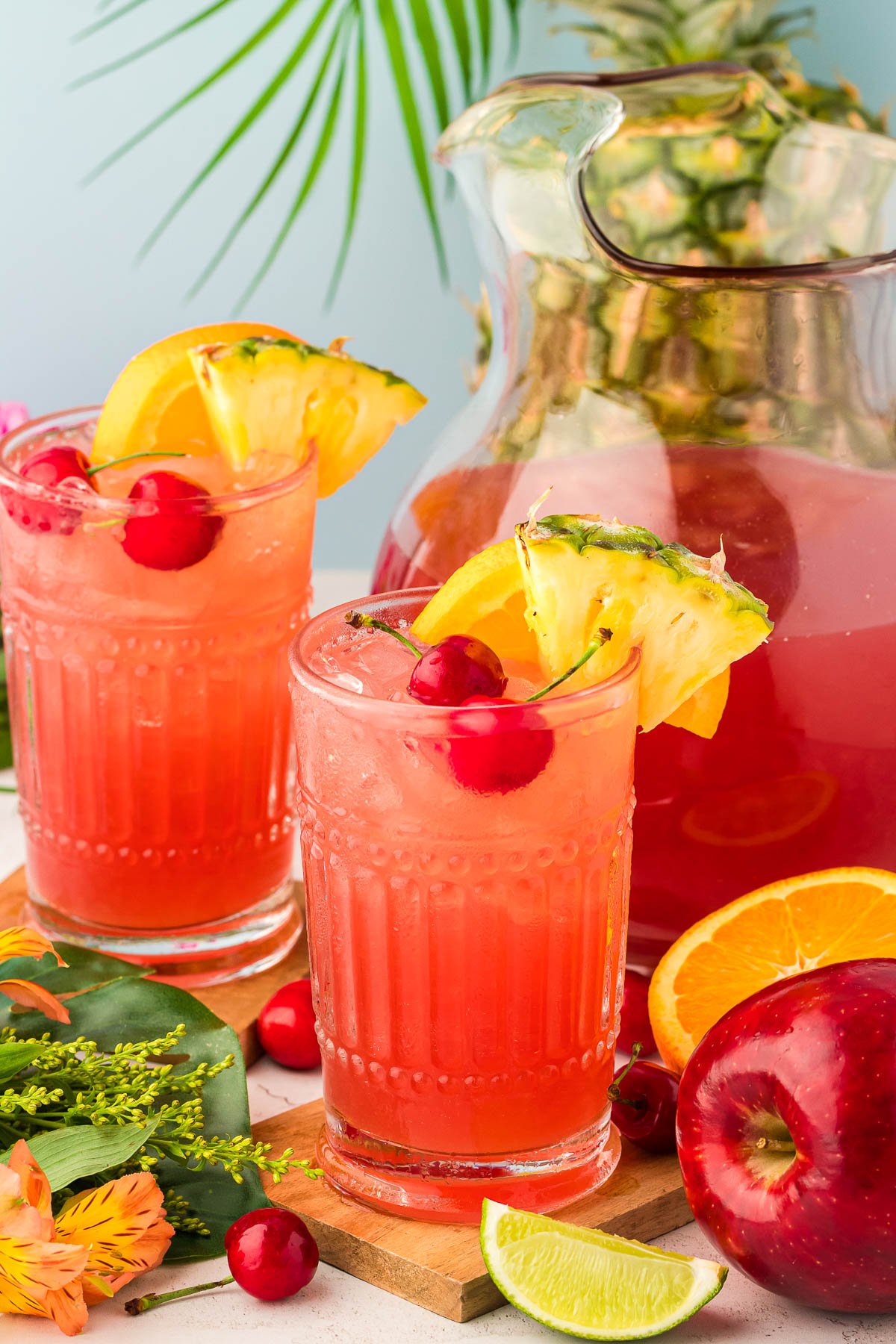 A glass of fruit punch on a wooden coaster with another glass, pitcher, and fruit in the background.