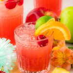 A glass of fruit punch on a wooden coaster with more glasses and fruit in the background.