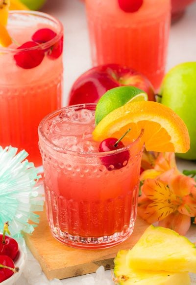 A glass of fruit punch on a wooden coaster with more glasses and fruit in the background.