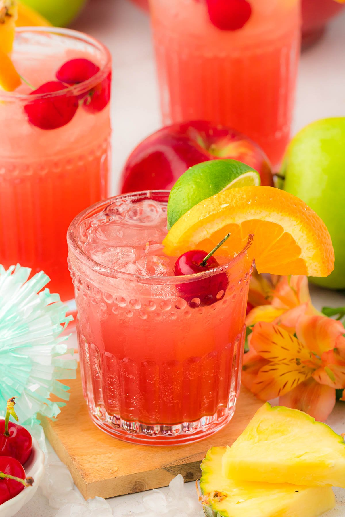 A glass of fruit punch on a wooden coaster with more glasses and fruit in the background.